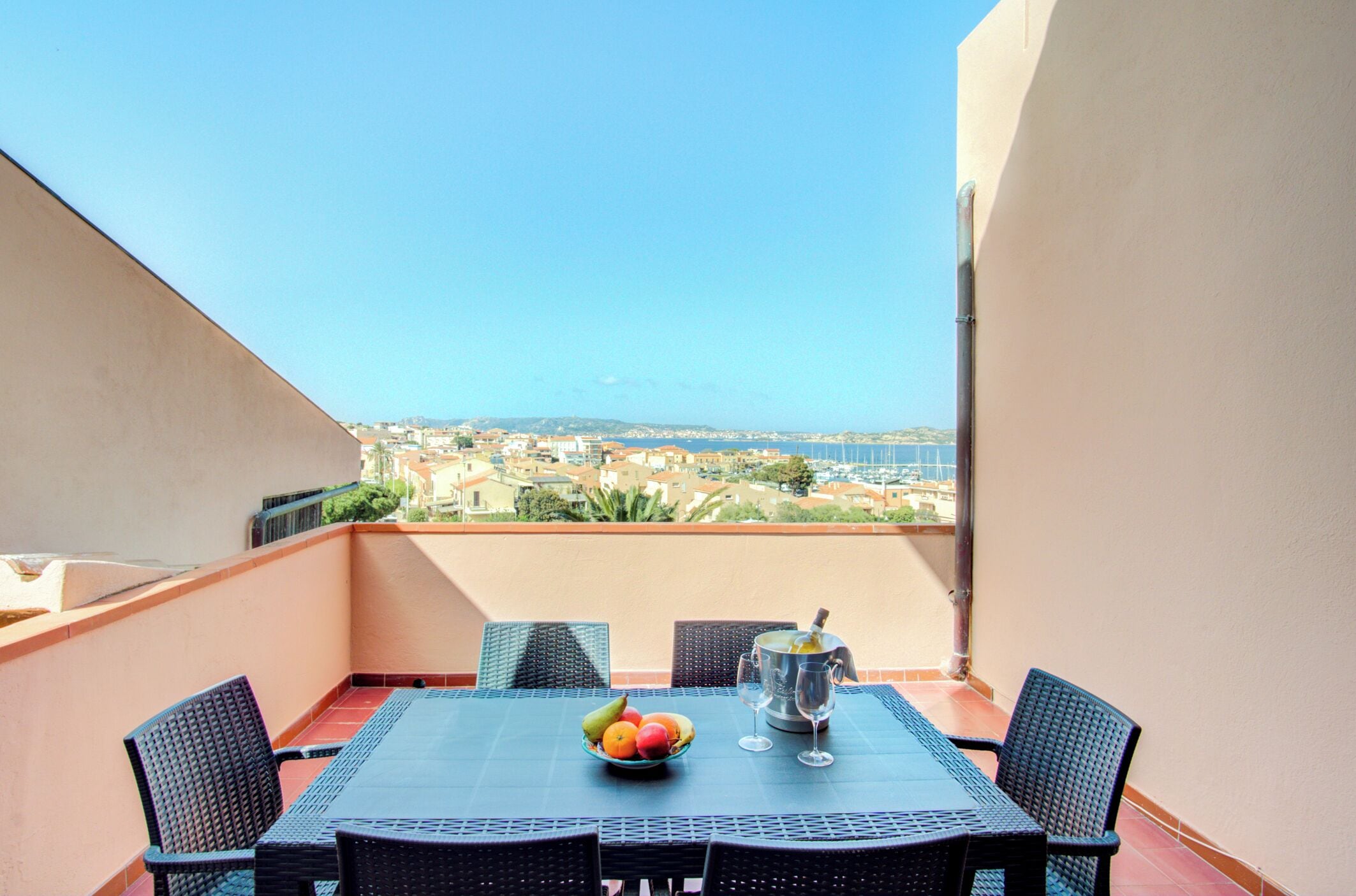 Un balcon avec une table dressée pour deux personnes, un panier de fruits et un verre à vin.