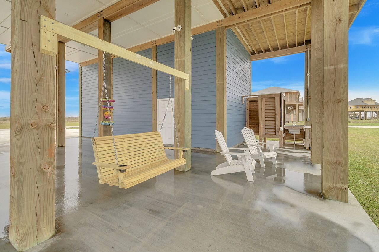 Un espace extérieur abrité, doté d’une balançoire en bois, de chaises Adirondack blanches et d’un sol en béton.