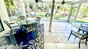 A covered patio with a glass table and chairs, a decorative rug, and a view of a garden.