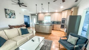 A modern kitchen with wooden cabinets, a central island, and built-in appliances. There is a dining area with a table and chairs, and a living room with a sofa and a coffee table.