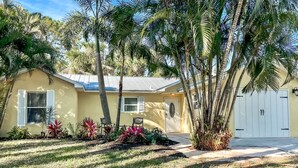 A yellow house with a white garage door, surrounded by palm trees and various plants.