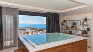 Una habitación moderna con un amplio jacuzzi de vidrio y vistas a una ciudad costera, además de un banco de madera.