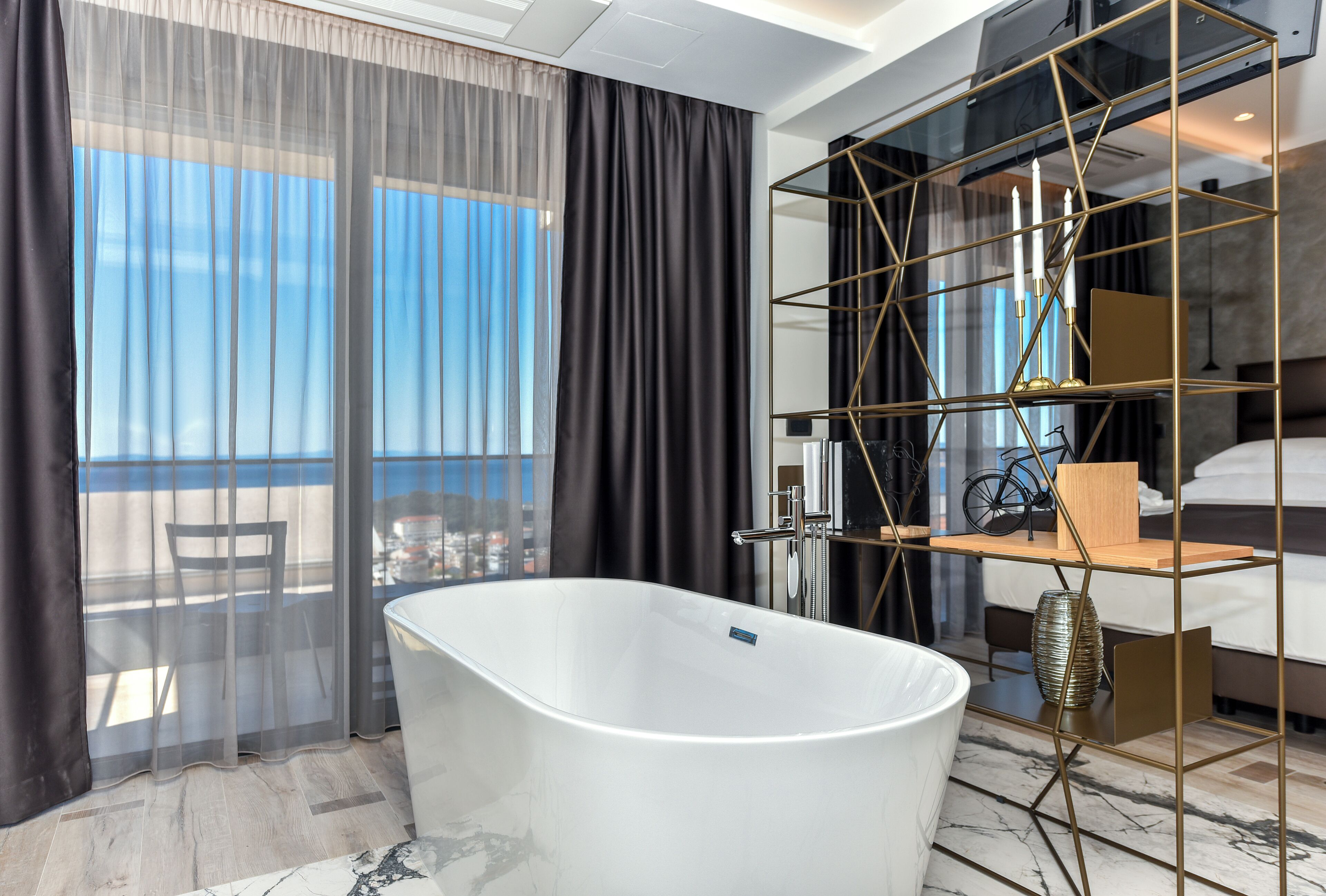 A modern bathroom with a freestanding bathtub, a glass shower enclosure, and a view of the city through sheer curtains.