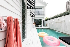 A modern outdoor pool area with a wooden deck, a white building with a balcony, and pink towels hanging on a railing. - Le Palms - A Beach House, Pool (Gold Coast)