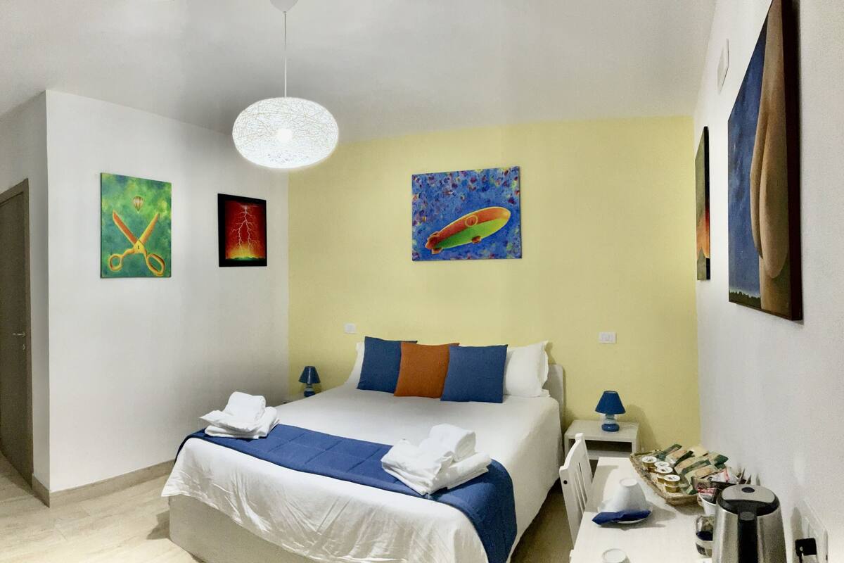 A hotel room with a bed, a desk, a coffee maker, and artwork on the walls.