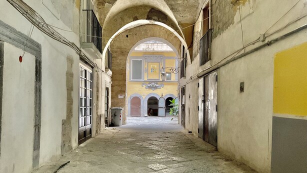A corridor with arched ceilings and a tiled floor, leading to a courtyard with a yellow building in the background. - A Room de Scugnizzi (Naples)