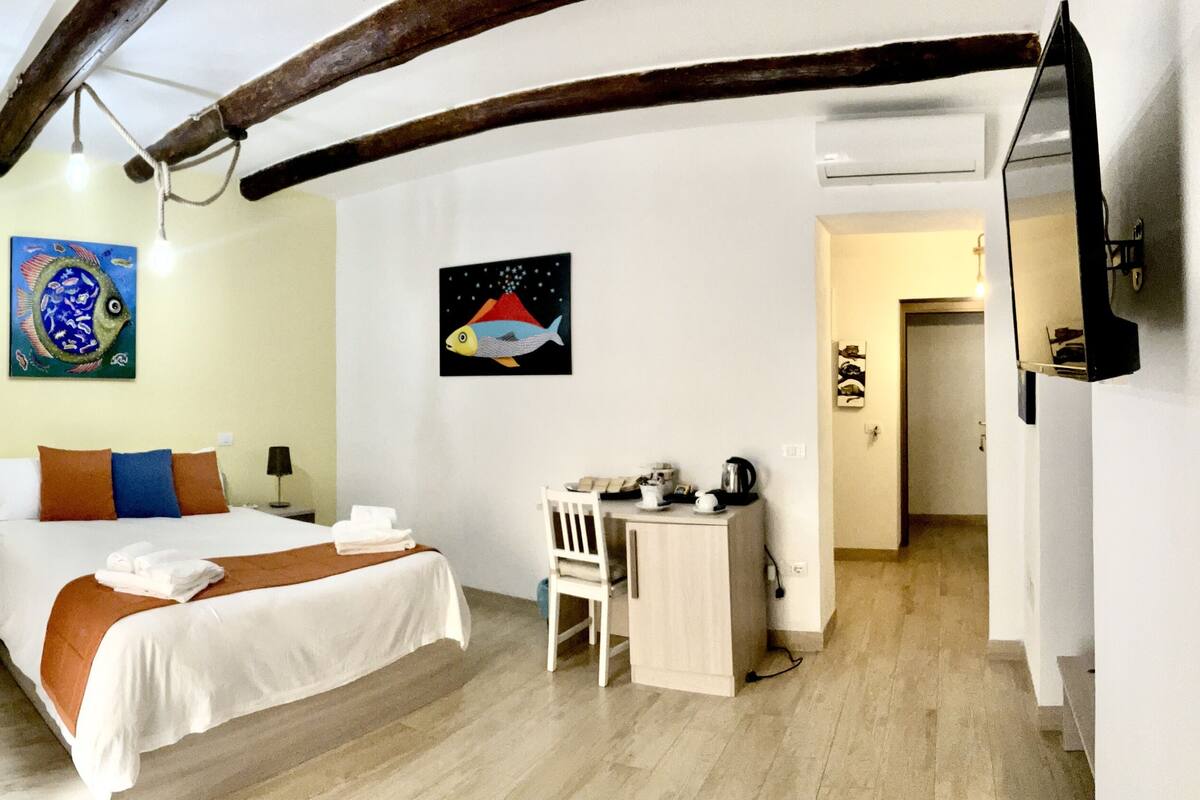 A hotel room with a bed, a desk, and a chair. There are paintings on the walls and a wooden ceiling.