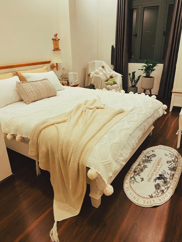 A neatly made bed with a white and cream-colored blanket, a bedside table with a lamp, a small potted plant, and a decorative plate on the floor. - A Nordic Design Apartment,close to the center, well prepared&super friendly host (Thành phố Đà Lạt)