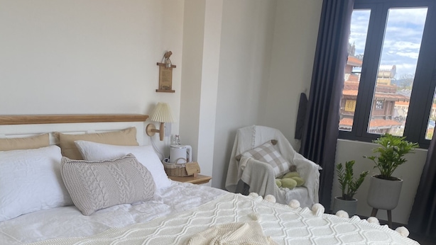A neatly made bed with a white knitted blanket and a pillow, a bedside table with a lamp, a small chair, and a view of buildings through a window. - A Nordic Design Apartment,close to the center, well prepared&super friendly host (Thành phố Đà Lạt)