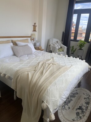 A neatly made bed with a white knitted blanket and a pillow, a bedside table with a lamp, a small chair, and a view of buildings through a window. - A Nordic Design Apartment,close to the center, well prepared&super friendly host (Thành phố Đà Lạt)