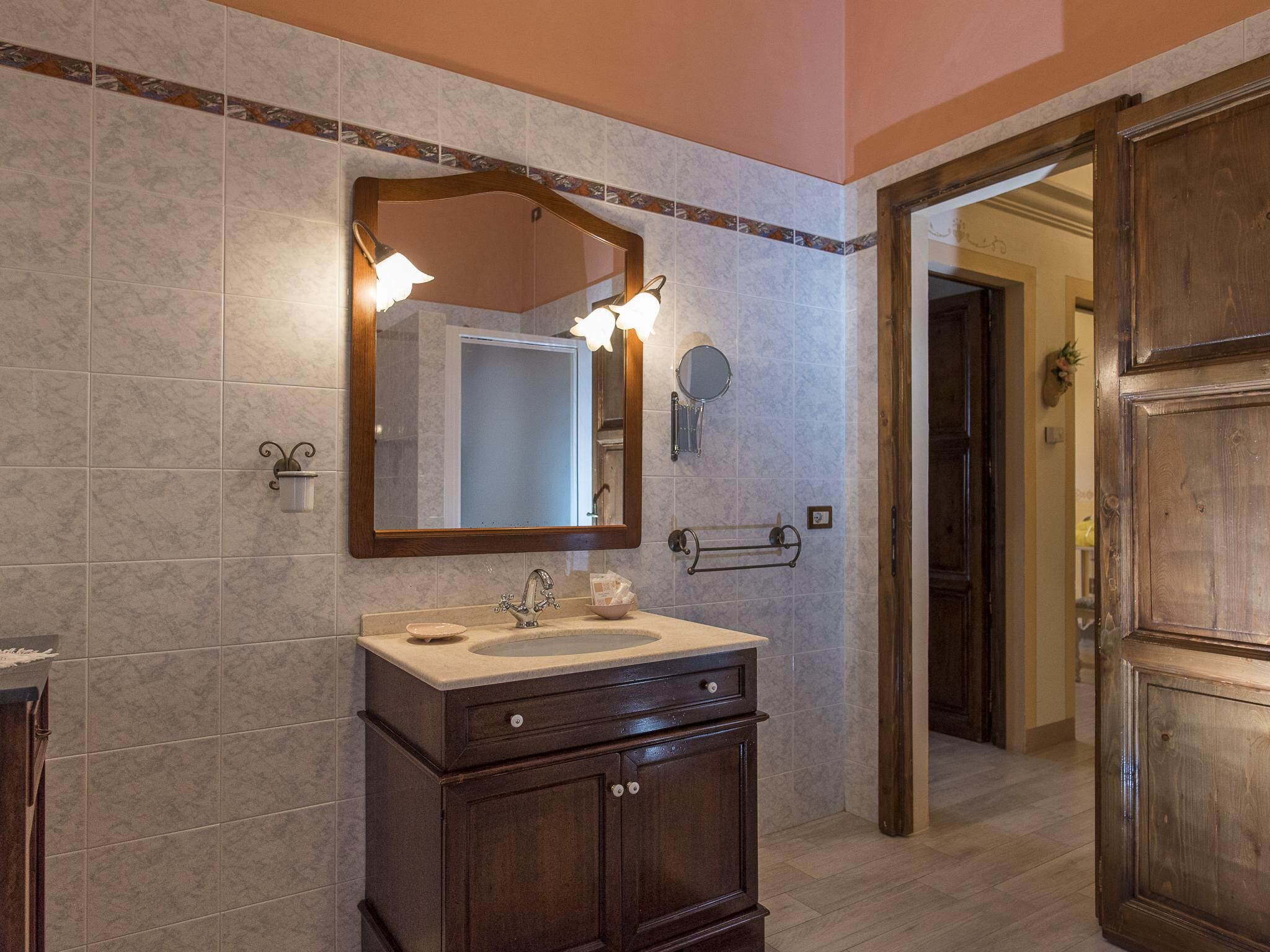 A bathroom with a marble-tiled wall, a wooden vanity with a sink, a large mirror, and a towel rack.