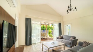 Una sala de estar moderna con televisor de pantalla plana, sofás grises, una mesa de centro y un amplio ventanal corredera que da a un patio con mobiliario exterior.
