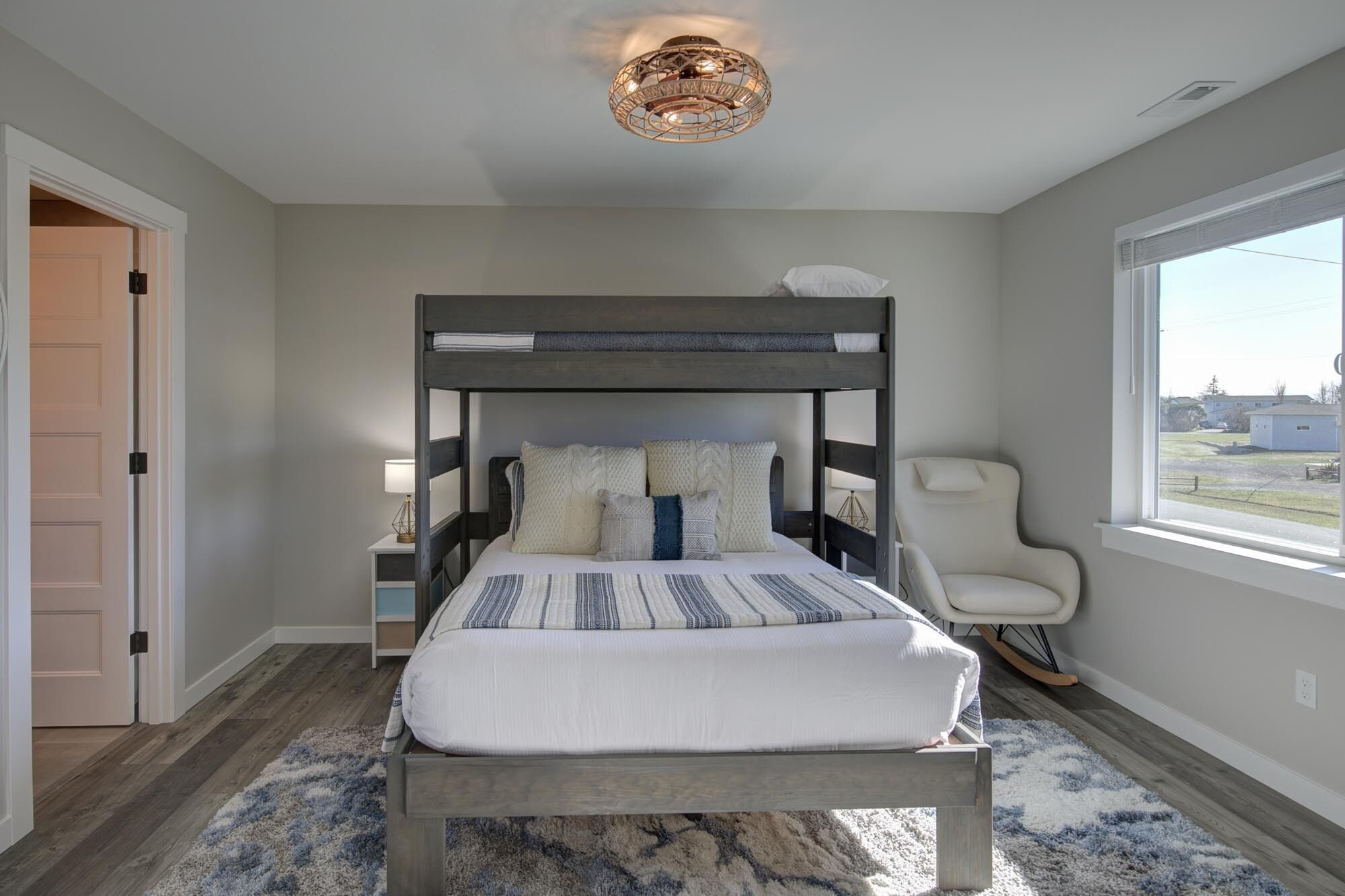 A bedroom with a bunk bed, a bedside table with a lamp, a rocking chair, and a large window offering a view.