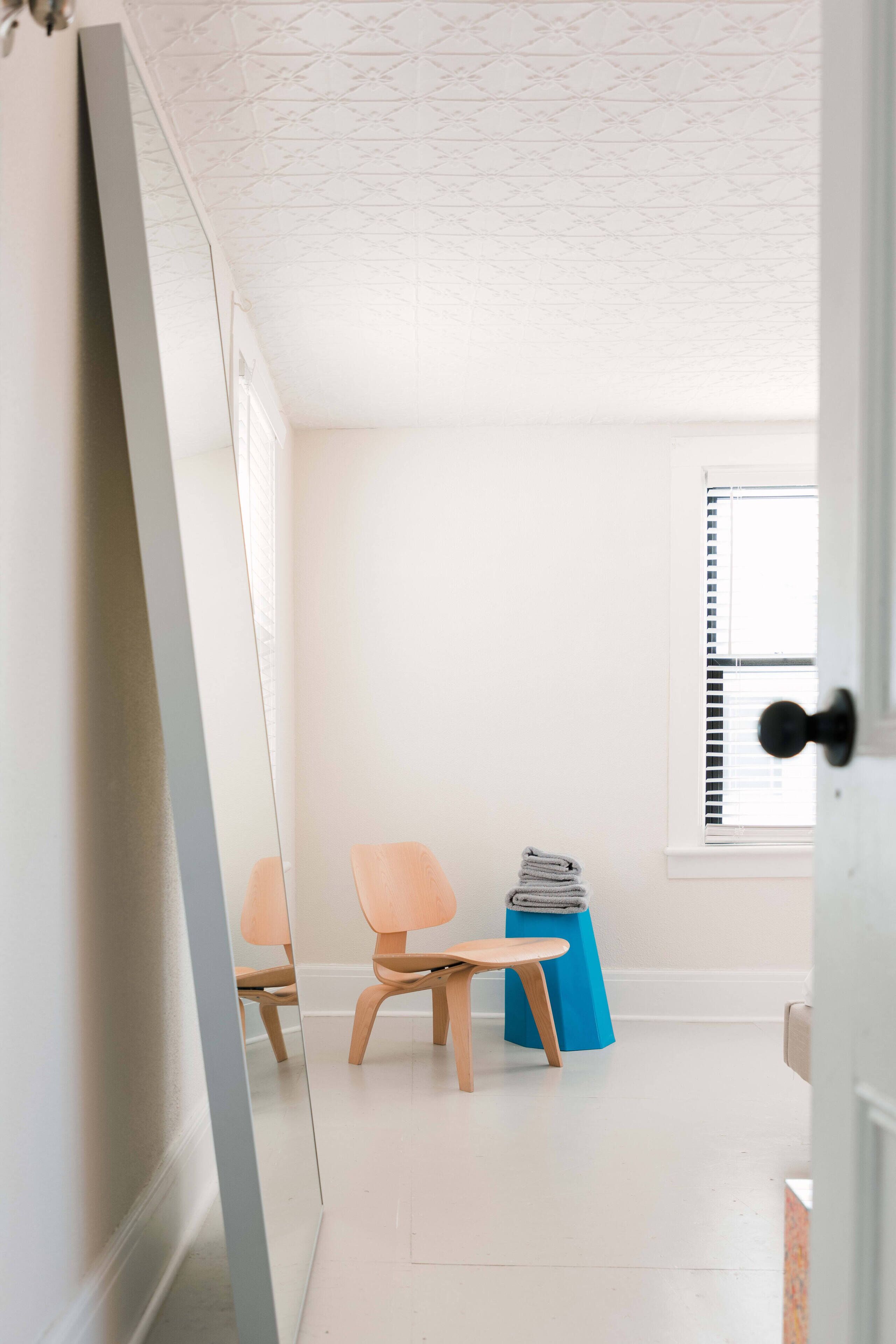 Una habitación con un espejo grande, dos sillas de madera, una mesita azul y una ventana con persianas.