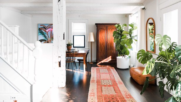 Un hall spacieux doté d’un plancher en bois, d’un tapis à motifs, d’un coin repas avec une table et des chaises, et de grandes plantes en pot.
