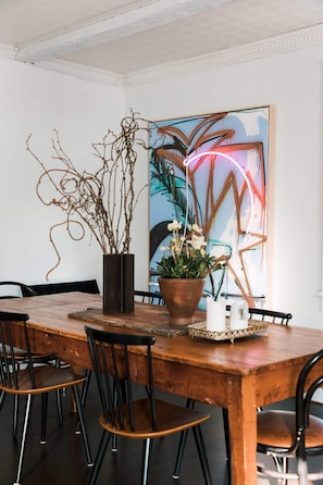 Uma sala de jantar com mesa de madeira, cadeiras pretas, um vaso com ramos e uma pintura abstrata colorida ao fundo.