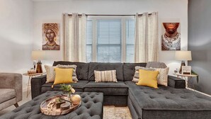 A modern living room with a sectional sofa, a round ottoman, and a coffee table. There are two side tables with lamps, a large window with blinds, and artwork on the walls.