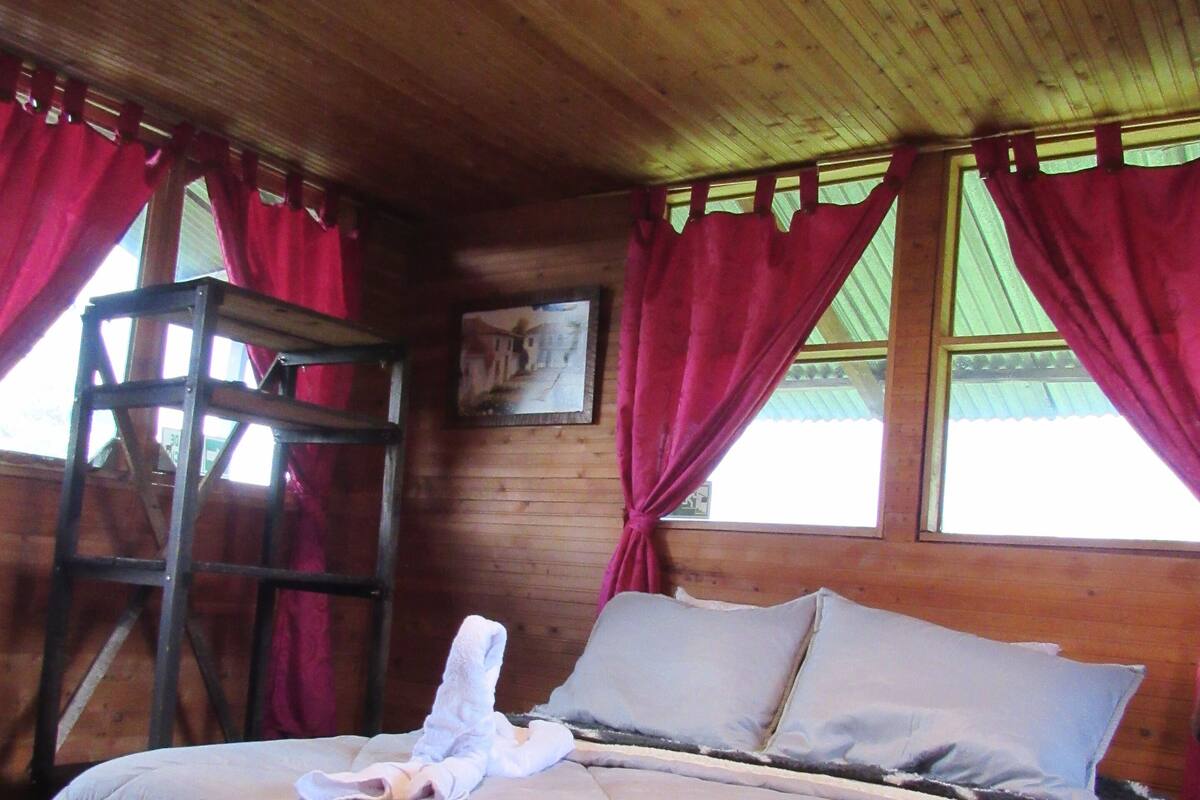 Ein Schlafzimmer einer Holzhütte mit einem Bett, roten Vorhängen, einer Leiter und einem Gemälde an der Wand.
