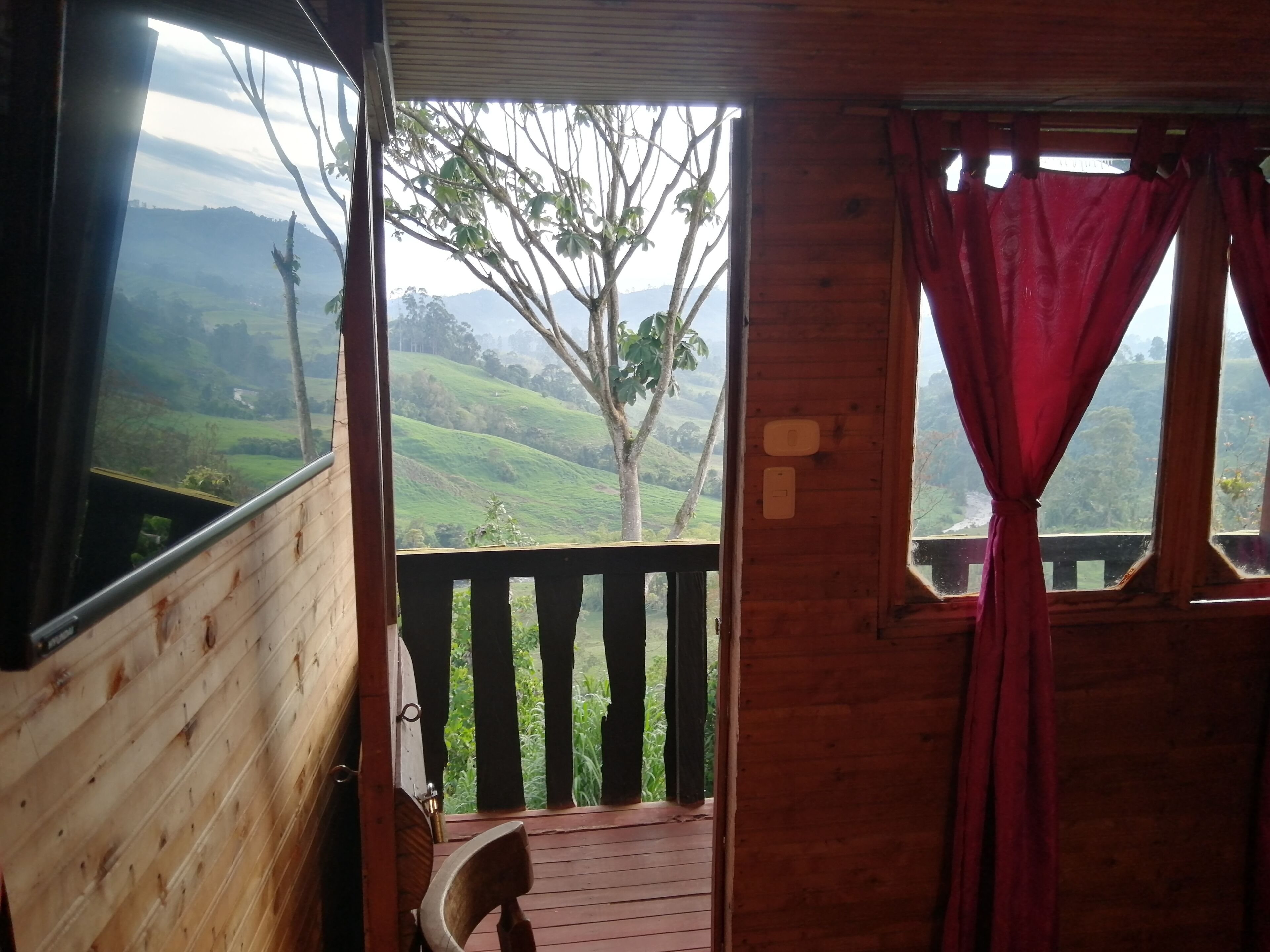 A wooden cabin with a balcony, a view of green hills, and a tree.