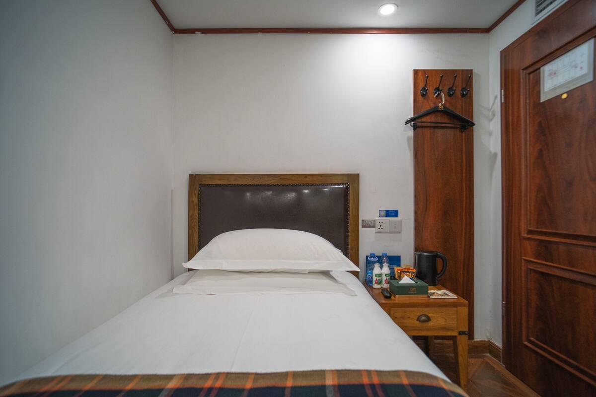 A hotel room with a single bed, a bedside table with a coffee maker, and a wooden door with a coat hanging on it.