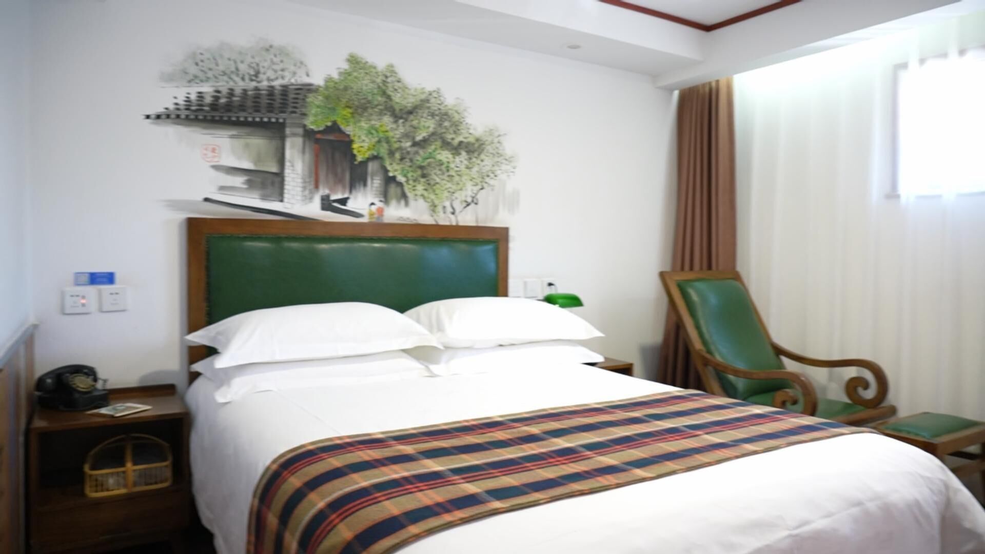 A hotel room with a bed, a nightstand, a chair, a plant, and a window with curtains.