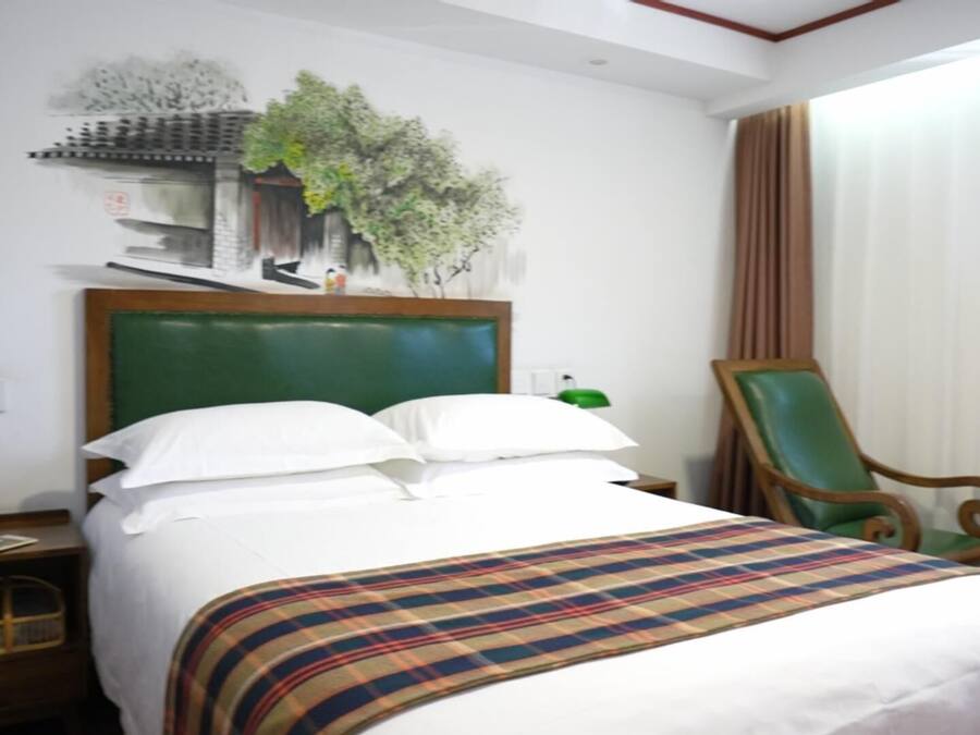 A hotel room with a bed, a nightstand, a chair, a plant, and a window with curtains.
