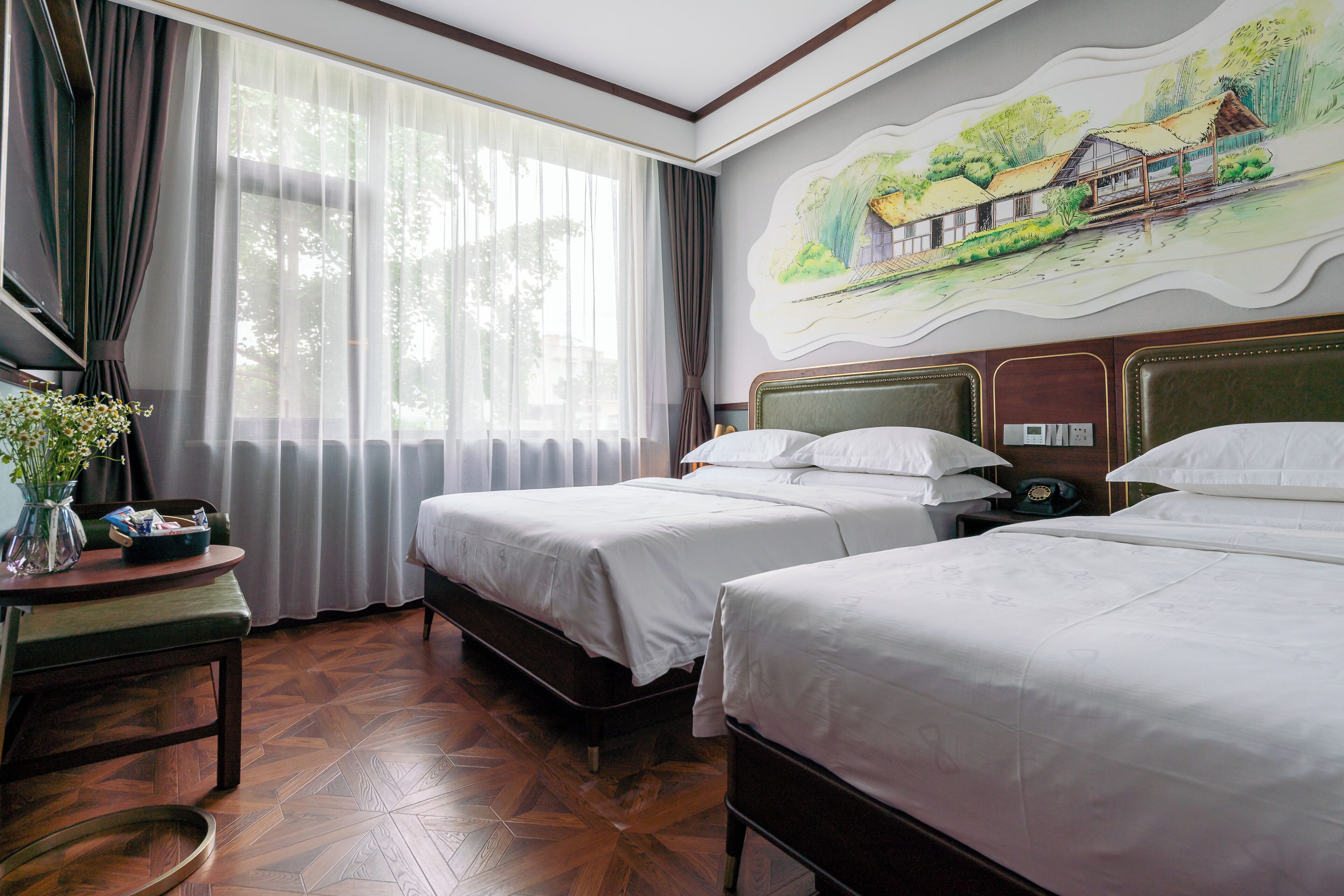 Habitació d'hotel amb dos llits, un escriptori amb un vas de flors, una finestra gran amb cortines i un mural a la paret.