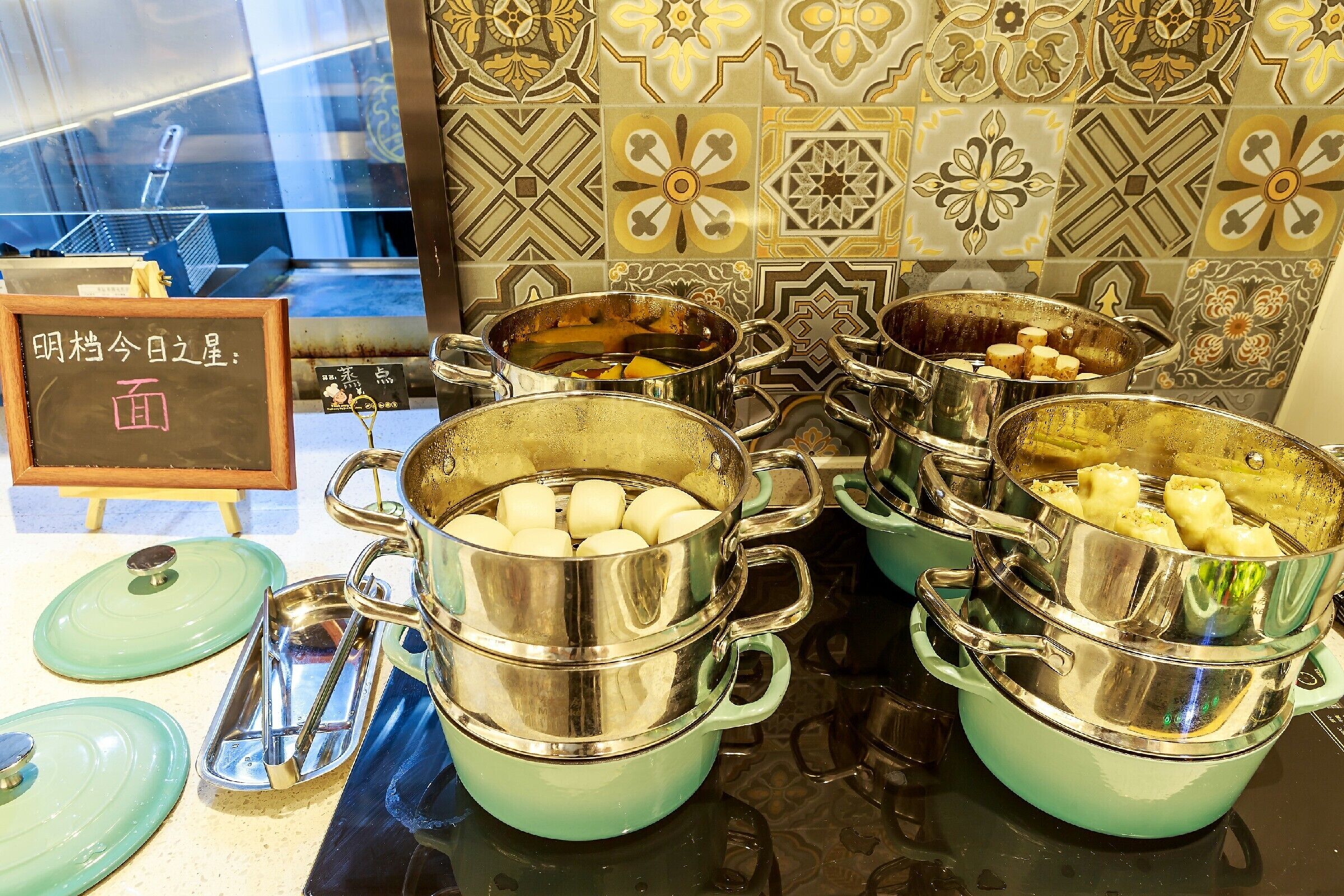 A kitchen area with multiple stainless steel pots and pans, some containing food items. There is a chalkboard with writing, a green plate, and a small sign with Japanese characters.
