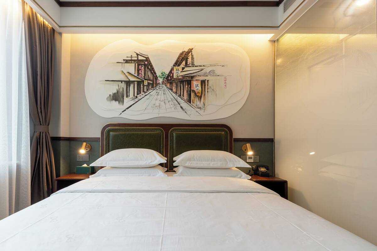 Une chambre d’hôtel avec un grand lit, deux tables de chevet et une œuvre d’art fixée au mur représentant une scène de rue.