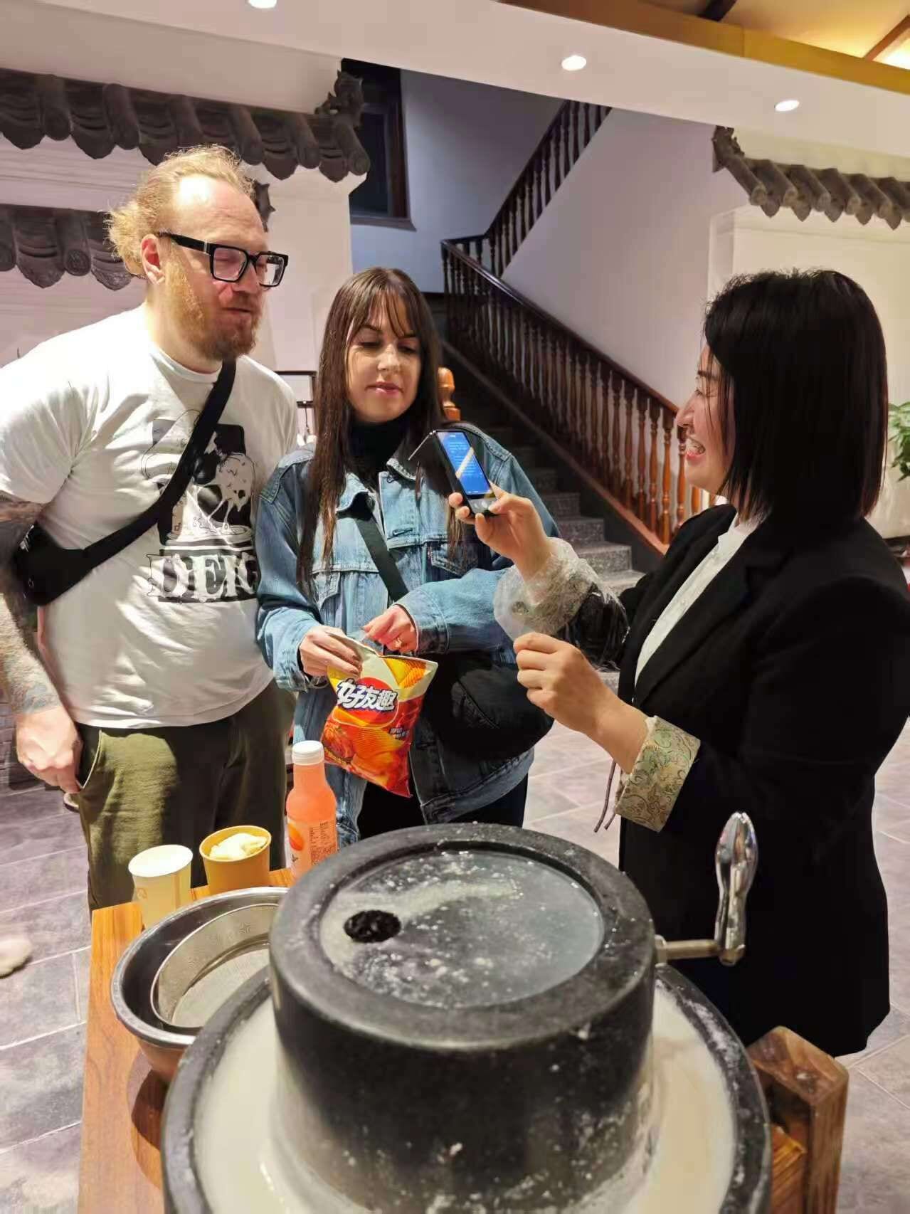 A group of people gathered around a table with a cake and a drink, in a room with a staircase and a tiled floor.