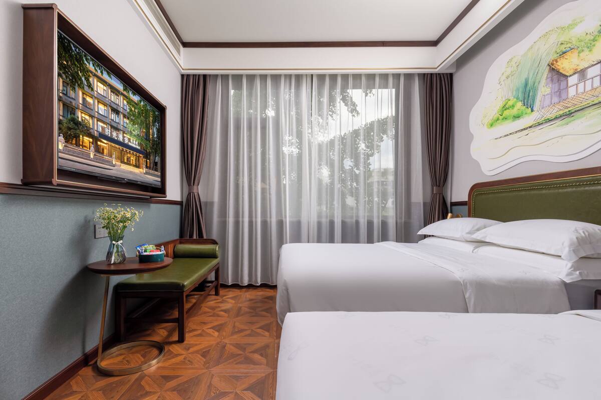 Une chambre d’hôtel avec deux lits, un bureau avec une chaise, une grande fenêtre avec des rideaux légers et un tableau encadré au mur.