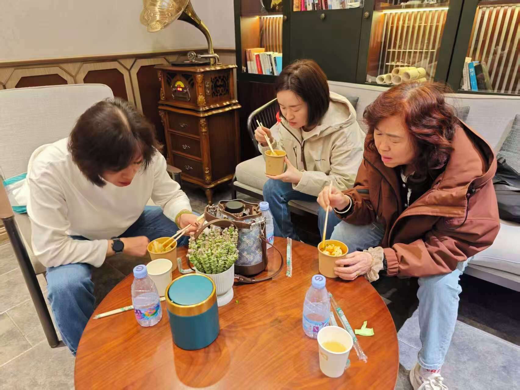 Un grupo de personas sentadas alrededor de una mesa de madera con comida y bebidas, en una habitación con una lámpara antigua y estanterías al fondo.