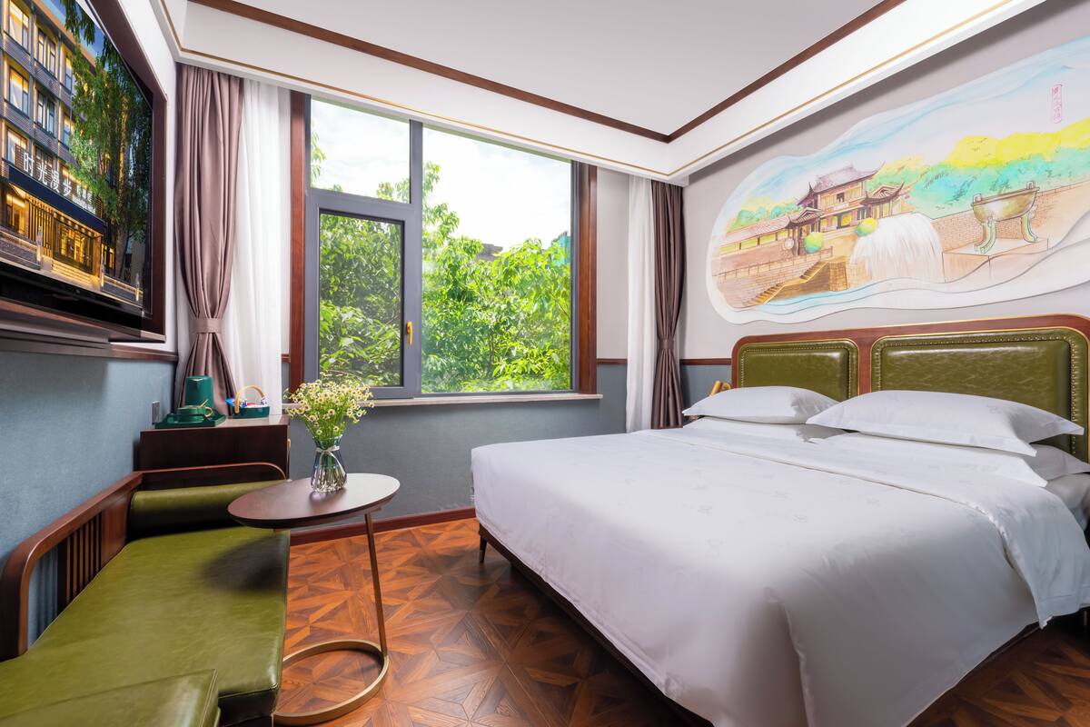 Une chambre d’hôtel avec un grand lit, une petite table ronde ornée d’un vase, un canapé vert et une fenêtre donnant sur un paysage verdoyant et un immeuble.