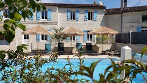 A poolside area with lounge chairs and thatched umbrellas, a stone building with blue shutters, and a clear blue pool. - Appart'é Nature l'ESCALE - SAINTES (Chaniers)