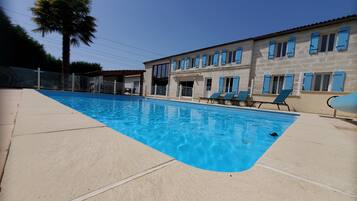 A swimming pool with clear blue water, surrounded by a concrete deck. There are several lounge chairs and a building with multiple windows and blue shutters in the background.