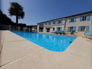 A swimming pool with clear blue water, surrounded by a concrete deck. There are several lounge chairs and a building with multiple windows and blue shutters in the background. - Appart'é Nature l'ESCALE - SAINTES (Chaniers)