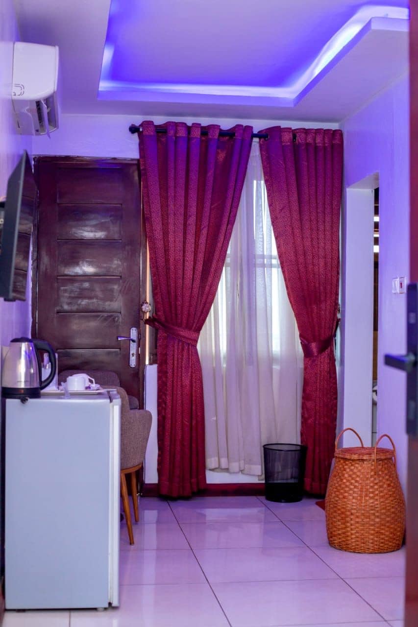 A compact living space with a kitchenette, a door with a handle, a small table with a chair, a television mounted on the wall, and a window with red curtains.