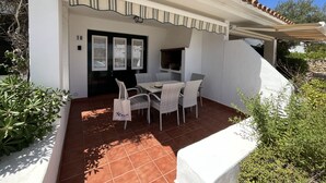 Un bâtiment blanc doté d’un patio carrelé, d’un espace de détente couvert avec des meubles en osier, et d’une porte noire avec des panneaux de verre.