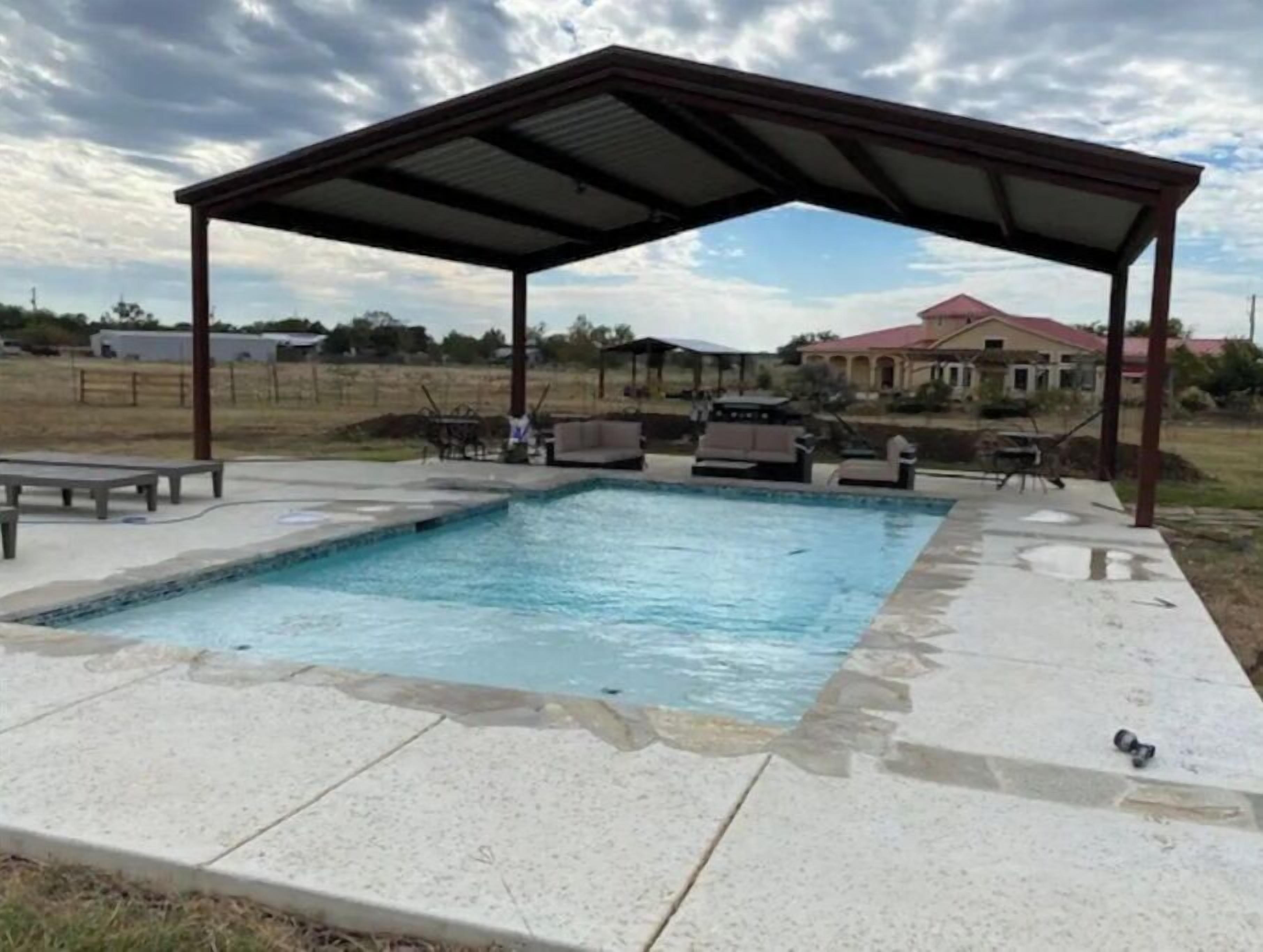 A swimming pool with a covered patio area, outdoor furniture, and a view of a house in the background.