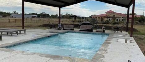 A swimming pool with a covered patio area, outdoor furniture, and a view of a house in the background.