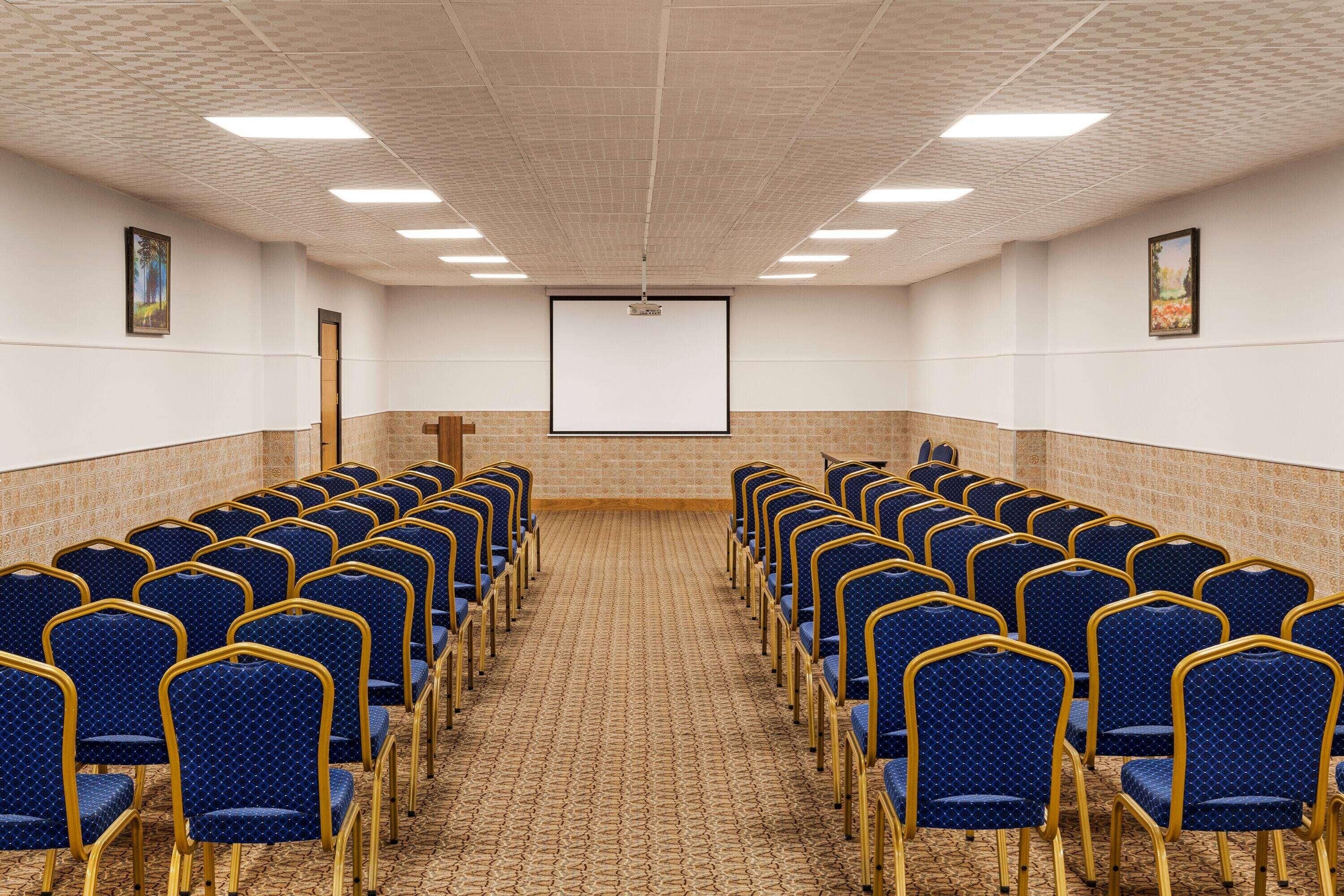 A conference room with rows of blue chairs, a projector screen, and a carpeted floor.