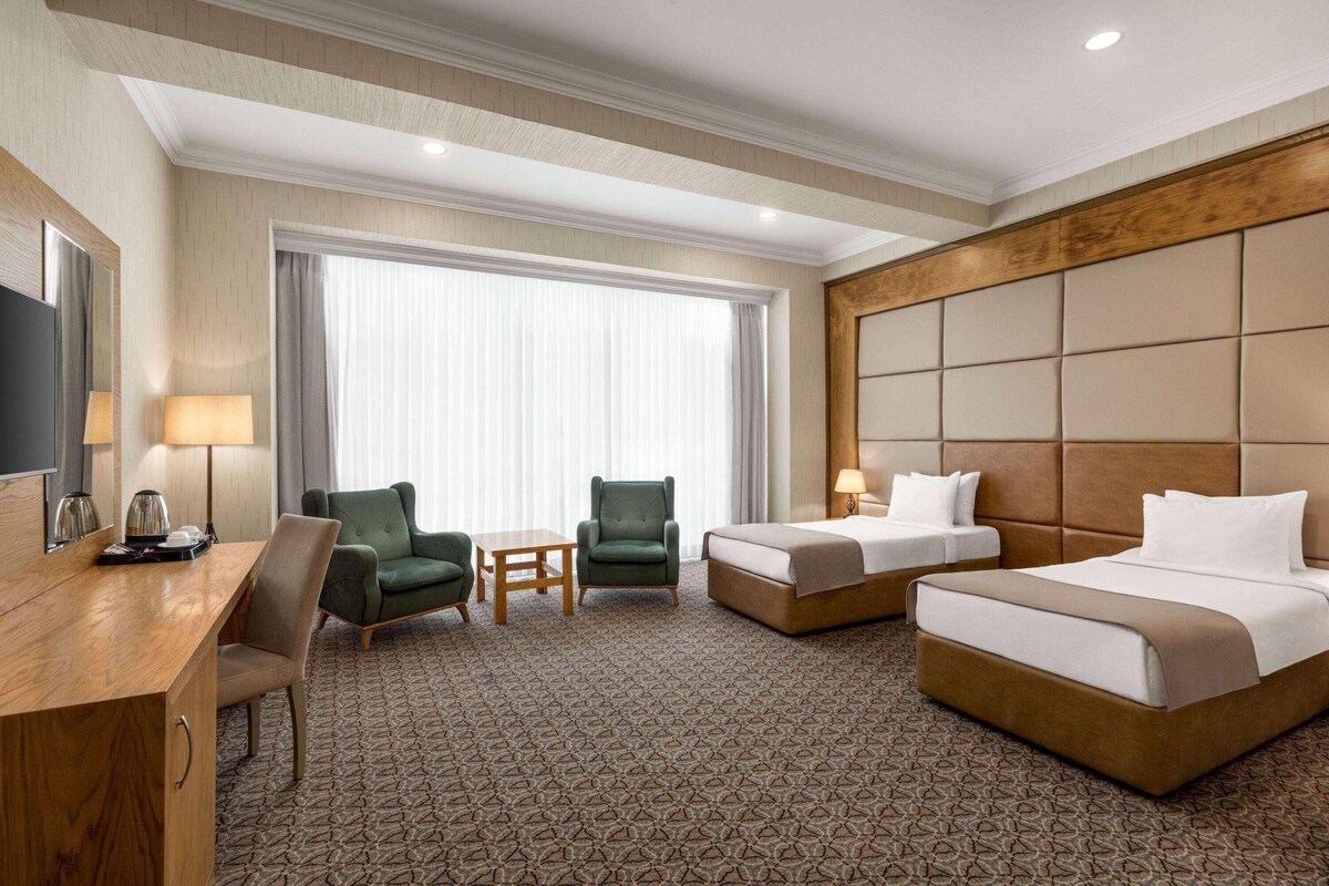 A hotel room with a bed, two armchairs, a desk, a TV, and a large window with curtains.