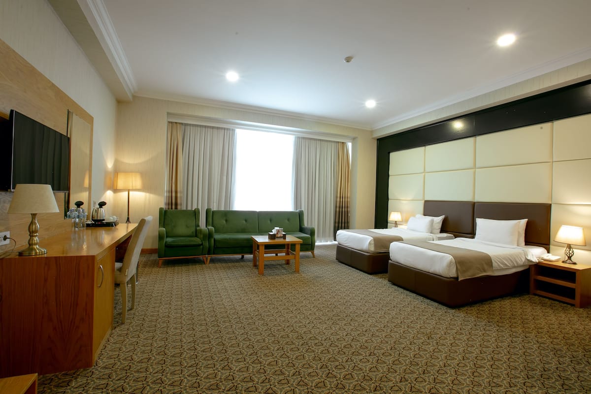A hotel room with a green sofa, a bed, a desk with a lamp, and a large window with curtains.