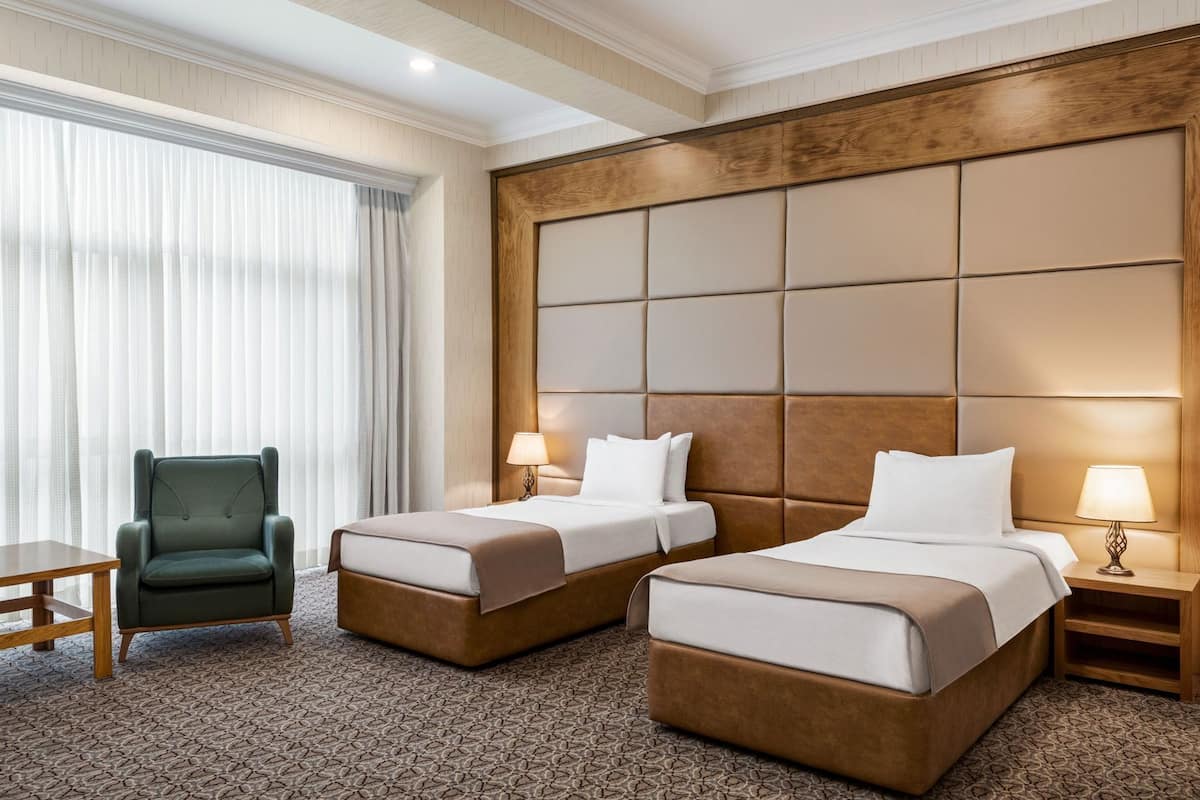 A hotel room with two beds, a chair, a table, and a large window with curtains.