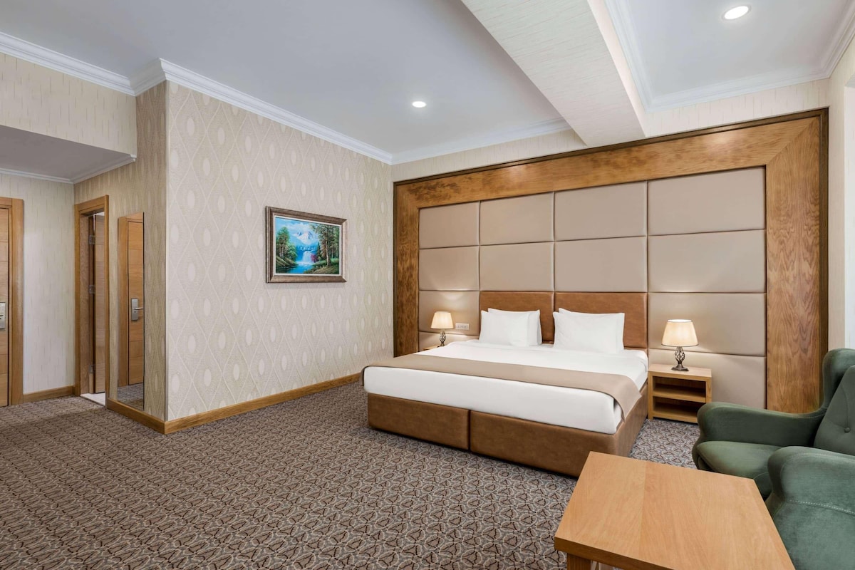 A hotel room with a large bed, two bedside tables with lamps, a green armchair, and a patterned carpet.