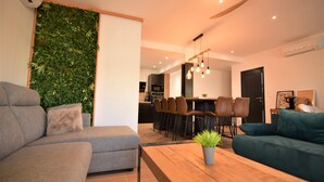 Ein modernes Wohnzimmer mit einem grauen Sofa, einem Holztisch und einer Zimmerpflanze. Es gibt einen Essbereich mit einem Holztisch und Stühlen, sowie eine Küche im Hintergrund.