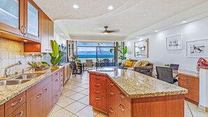 A spacious kitchen with granite countertops, wooden cabinets, and a large island. There is a dining area with chairs and a table, and a balcony with a view of the ocean. - Oceanfront | Walk to Beach  | Hale Mahina B305 | (Lahaina)