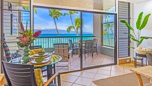 A balcony with a glass door, a dining table with chairs, and a view of the ocean and palm trees. - Oceanfront | Walk to Beach  | Hale Mahina B305 | (Lahaina)
