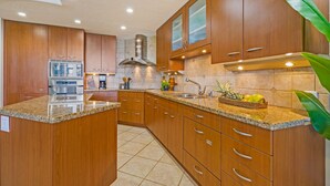 A modern kitchen with wooden cabinets, granite countertops, stainless steel appliances, and a central island. - Oceanfront | Walk to Beach  | Hale Mahina B305 | (Lahaina)