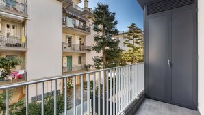 Un balcón con una puerta gris, barandilla metálica y vistas a edificios y árboles cercanos.